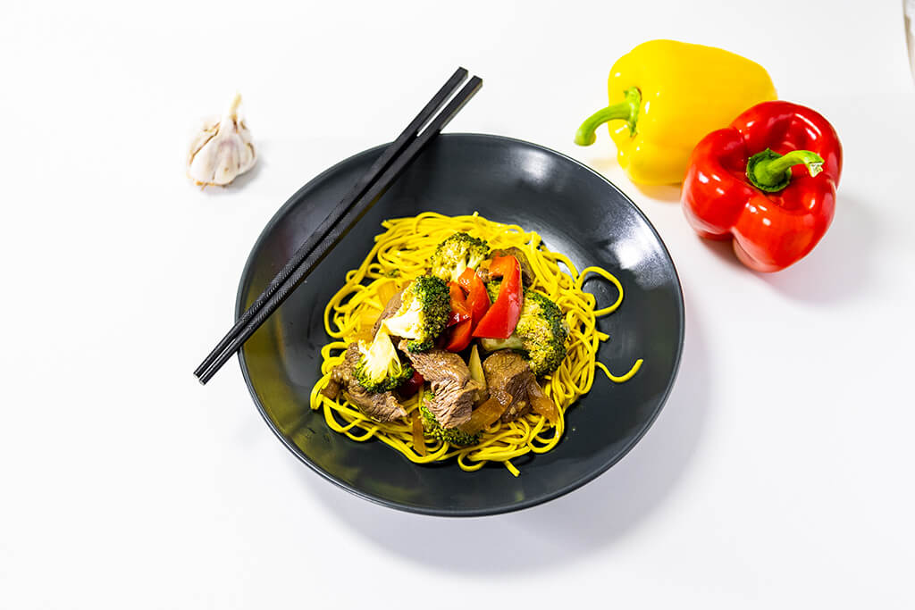 Beef Stir Fry in a black bowl with chop sticks and garlic/peppers surorunding